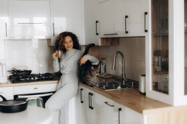 Modern beyaz bir mutfakta elinde kahve fincanı olan bir kadın. Tezgahta kedisiyle konuşurken gülümsüyor. Sahne konfor, konfor ve günlük hayat anlarını aktarır..