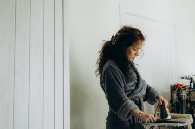 Pofuduk bornoz giyen bir kadın, rahat ve aydınlık bir odada kıyafetlerini ütülüyor ve işine odaklanıyor. Atmosfer konforu ve rutini yansıtıyor. Minimalist iç tasarım ve sade detaylarla..