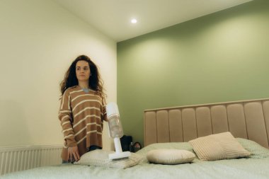 Modern bir yatak odası ortamında elinde kablosuz elektrik süpürgesi tutan bir kadın. Odada yeşil ve bej renkler, minimalist ve rahat bir ortamda rahatlık ve temizlik var..