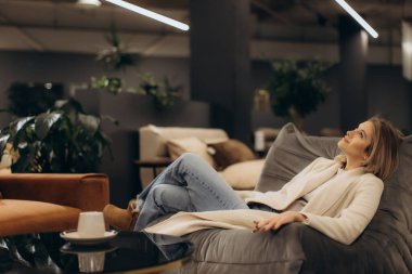 Şık bir oturma odasındaki yumuşak kanepeye yaslanan bir kadının huzur, rahatlama ve modern iç mekan estetiğini anımsattığı sakin bir sahne..