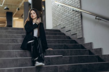 Ceket giyen kendine güvenen bir kadın merdivenlerde düşünceli bir şekilde oturur çağdaş bir ortamda, moda ve tarz göstererek. İmaj, kurumsal veya kentsel bir ortamda gelişmişliği vurgulayarak profesyonelliği ve rahatlığı iletir..