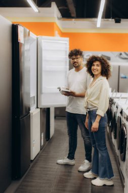 Gülümseyen bir çift, elektronik mağazasında alışveriş yaparken buzdolaplarını keşfeder ve evleri için seçenekleri tartışır..