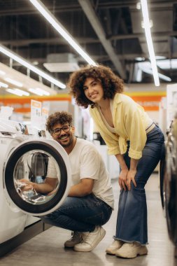 Neşeli bir çift modern bir elektronik mağazasında alışveriş yaparken bir çamaşır makinesini inceliyor. Parlak ortam ve mutlu ifadeler yeni ev aletleri seçmenin heyecanını vurguluyor.