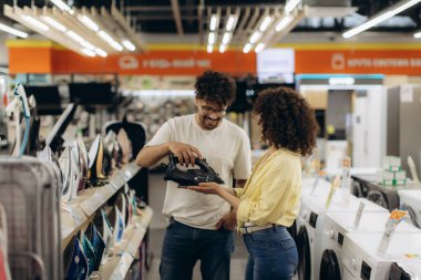 Modern bir mağazadaki genç bir çift, çeşitli elektronik aletler arasında satın alma kararlarını verirken ütüyü inceliyor..