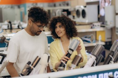 Neşeli bir çift modern bir elektronik mağazasında çeşitli saç şekillendirme aletleri keşfediyor. Çift saç maşası, saç kurutma makinesi, saç düzleştirici alır.