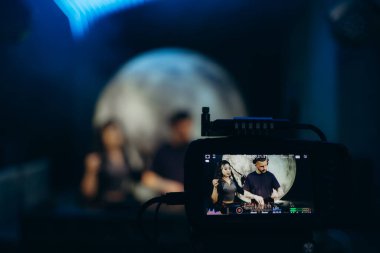 Yaratıcı bir proje üzerinde çalışan bir takımı yakalayan bir video kameraya odaklanan sanatsal bir düzenek. Arka planda, vizyon sahibi ve sanatsal bir atmosfer yaratan, yumuşak, parlak bir ortam yer alıyor..