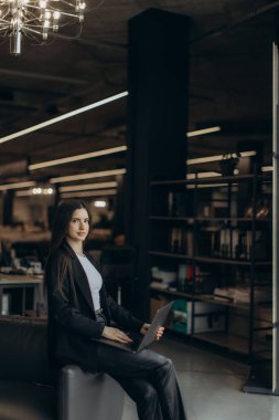 Çağdaş bir ofiste otururken dizüstü bilgisayarda çalışan bir iş kadını profesyonelliği ve modern çalışma ortamlarını gösteriyor..