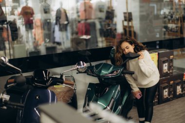 Şık bir genç kadın modern bir elektronik mağazasında mobiletini dikkatle inceliyor. Sahne, tüketici karar verme mekanizmasını, merakı ve yeni teknolojinin canlı şehir ortamında keşfedilmesini betimliyor..