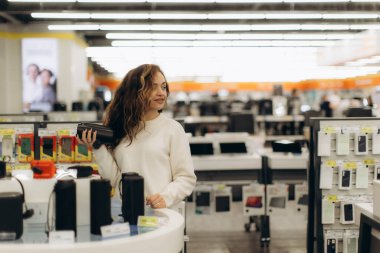 Genç bir kadın, taşınabilir cihazlara ve yenilikçi cihazlara ilgi gösteren parlak ışıklı bir teknoloji mağazasında elektronik aletleri keşfediyor..