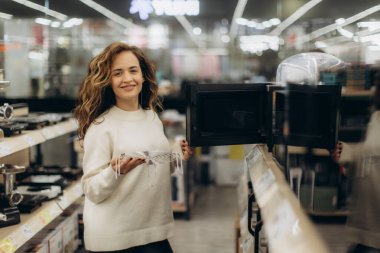 Genç bir kadın bir elektronik mağazasında duruyor, mikrodalga fırın seçerken gülümsüyor. Parlak ve modern alışveriş ortamı.