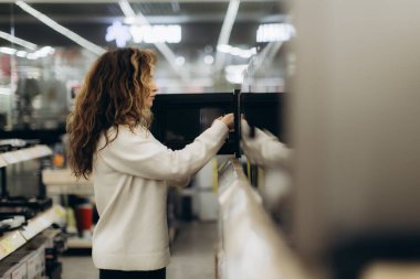 Kadın modern perakende satış alanında elektronik aletleri keşfediyor, özenle seçilmiş ürünler sergiliyor. Modern bir mağaza ortamında eğlence aktivitesi, alışveriş odağı ve teknolojiyle ilgili karar verme mekanizmasını yakalar.
