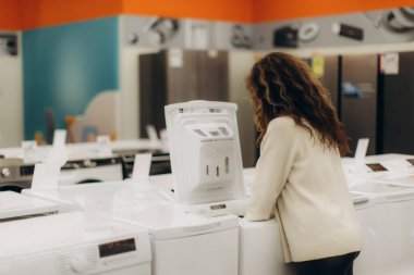 Genç bir kadın, modern bir elektronik mağazasında çeşitli aletleri inceleyip alışveriş seçimleri yapıyor. Görüntü çağdaş bir ortam yakalıyor ve tüketim, alışveriş ve teknoloji temalarını keşfediyor..
