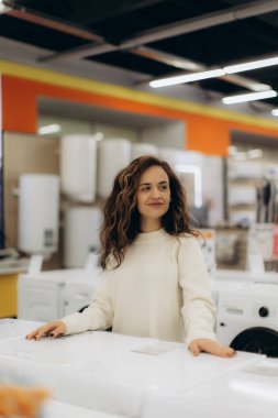 Genç bir kadın, bir elektronik mağazasında sergilenen çeşitli ev aletlerini araştırıyor, araştırma yaparken merak ve ilgi gösteriyor. Modern ışıklandırma ve görüntüleme alışveriş deneyimini artırır.