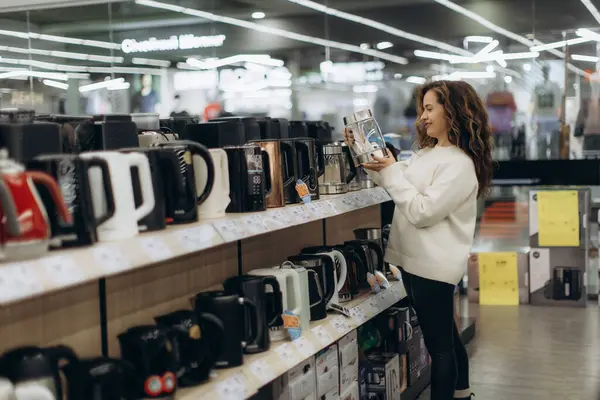 İyi aydınlatılmış bir elektronik mağazasında elektrikli çaydanlık arayan bir kadın. Günlük giysiler giyer. Raftaki bir ürünü değerlendirirken merakını ve düşüncelerini yansıtır..