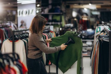 Süslü bir giyim mağazasında yeşil bir kazağın üzerinde vapur kullanan bir kadın. Görüntü düzgün ve profesyonel bir perakende satış ortamını tasvir ediyor..