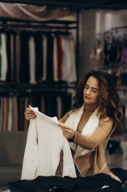Bir kadın şık bir giyim mağazasında beyaz bir bluzu inceliyor, modaya uygun kıyafetler almaktan zevk alıyor..