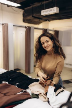 Bir giyim mağazasında katlanmış kıyafetlerin yanında duran neşeli bir kadın, sıcak bir şekilde gülümsüyor. Çevrede, arka planda uygun odalar bulunan ve keyifli bir alışveriş deneyimi sergileyen sıradan bir alışveriş ortamı yer alıyor..