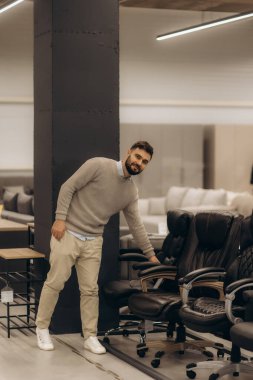 Günlük kıyafet giyen bir adam, bir alışveriş deneyiminde ürün kalitesi ve tasarımı vurgulayarak çağdaş bir perakende galeride ofis sandalyeleri koleksiyonunu teftiş eder..