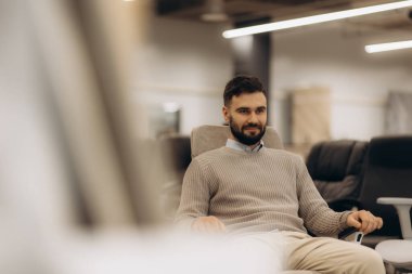 Neşeli bir adam modern bir koltukta rahatça oturuyor, kendine güvenerek gülümsüyor, şık bir sergi salonuyla çevrili. Ortam hoş karşılanıyor ve ticari mobilya ortamında rahatlama ve memnuniyet gösteriyor..