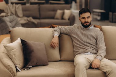 Sıradan bir süveter giyen sakallı bir adam, bir mobilya galerisindeki rahat bir bej kanepeye oturmuş, odanın sofistike iç dekorunun arasında rahat ve huzurlu bir havayı yansıtıyor..
