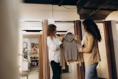 İki kadın şık bir butik ortamında kıyafet seçerken sohbet ediyorlar..