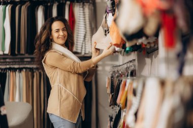 Neşeli bir kadın moda mağazasında kıyafetleri karıştırıyor, alışveriş deneyiminden zevk alıyor. Düzenli bir raftan canlı bir perakende atmosfer sergileyerek eşyalar seçerken gülümsüyor..