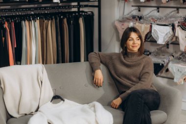 Modern bir giyim mağazasında şık bir kadın rahat bir koltukta oturuyor, giysi, iç çamaşırı, vitrin alışverişi ve yaşam tarzı estetiği gibi çeşitli moda eşyalarıyla çevrili..