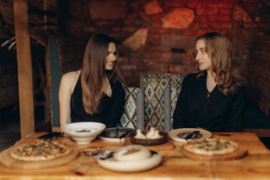 Ilık bir restoranda yemek yiyen iki kadın, gülümsemelerini ve sohbetlerini masada samimi bir ortam ve lezzetli yemeklerle paylaşıyorlar..