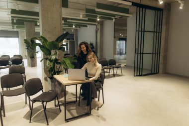 İki kadın, geniş ve modern bir ofis ortamında birlikte çalışıyor. Arka planda düzgün sıralanmış sandalyeler ve uzayın estetiğini geliştiren bitkiler var..