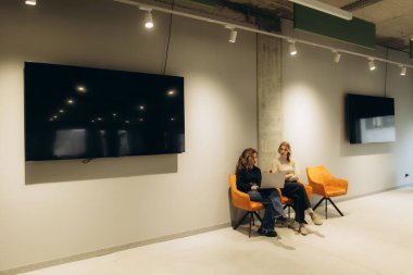 Çağdaş bir ofis salonunda oturan iki kadın, turuncu sandalyeler ve duvarlara monte edilmiş ekranlar da dahil olmak üzere modern dekorlarla çevrili bir dizüstü bilgisayarın üzerinde tartışıyorlar. İşbirlikçi ve profesyonel bir ortam yaratıyorlar..
