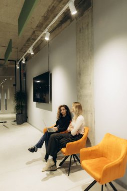 Modern bir salonda oturmuş fikirleri tartışan iki kadın, dizüstü bilgisayarları ve parlak dekorlarıyla, işbirlikçi bir profesyonel ortamı vurguluyorlar..