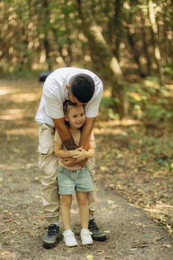 Bir babanın güneşli bir orman patikasını keşfederken kızını kucaklaması, sevgiyi ve bağlantıyı sembolize eder..