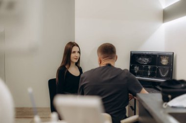 Dişçi hastalarla tedavi seçeneklerini tartışıyor, modern bir klinikteki bilgisayar ekranında diş röntgenlerini analiz ediyor. Sağlık hizmetlerinde diş sağlığı, profesyonellik ve teknolojinin vurgulanması.