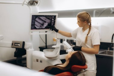 Modern bir klinikte diş röntgeni sonuçlarını gösteren beyaz üniformalı profesyonel bir dişçi. Görüntü diş sağlığı, hasta iletişimi ve diş hekimliğindeki sağlık uzmanlığını temsil etmektedir..