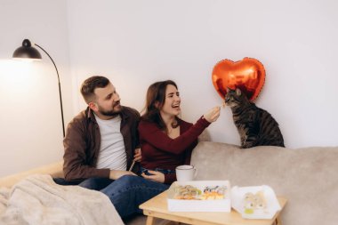 Neşeli bir çift birlikte vakit geçirir, kedilerini kalp şeklinde bir balonun yanında neşeyle beslerler ve sıcak ve sıcak bir ev ortamında yemeklerini yerler..
