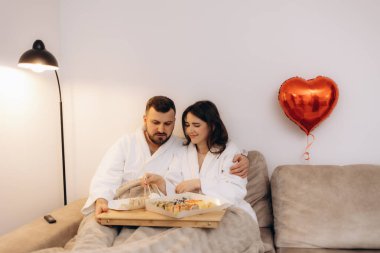 Bir çift, ahşap bir tepsi suşiyle koltukta rahatlıyor, romantik ve samimi bir ortam yaratıyor, kalp şeklinde bir balon ve yumuşak, samimi bir ortam tarafından vurgulanıyor..