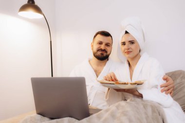 Bornoz giyen bir çift evde vakit geçirmekten, atıştırmalıkları paylaşmaktan ve sıcak, rahat bir atmosferde dizüstü bilgisayar kullanmaktan keyif alır..