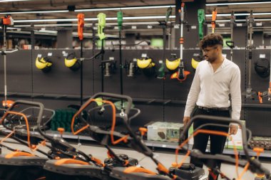 Bir adam, çim biçme makinesi ve başlık gibi seçenekleri düşünerek bir perakende mağazasında bahçe ekipmanlarını inceliyor. Mağaza, çeşitli alet ve aksesuarlarla aydınlık ve iyi organize edilmiş..