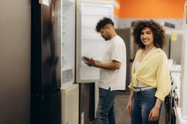 Neşeli bir çift, bir elektronik mağazasından alışveriş yaparken çeşitli buzdolabı seçeneklerini araştırıyor. Partneri buzdolabını yakından incelerken kadın kameraya sıcak bir şekilde gülümsüyor..