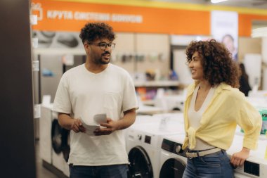 Çağdaş bir elektronik galeride çamaşırhane için seçenekleri araştıran neşeli bir çift, özellikleri ve tasarımları faiziyle tartışıyor..