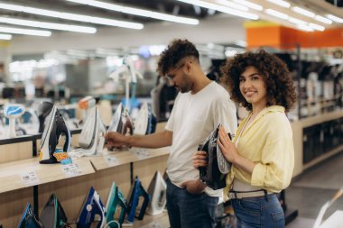 A cheerful young couple examines irons while shopping in a brightly lit electronics store.