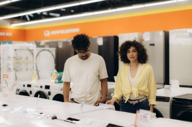 Bir çift çağdaş bir elektronik mağazasını araştırıyor, aletleri inceliyor ve satın almak için özelliklerini değerlendiriyor..