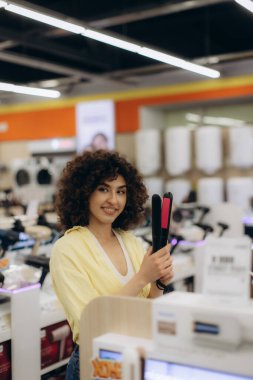 Elektronik mağazasında alışveriş yapan bir kadın elinde neşeli bir ifadeyle saç düzleştiricisi tutuyor ve sergiliyor. Düzenli raflarda çeşitli aletlerle modern perakende satış ortamı.
