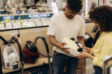 Genç bir çift bir elektronik mağazasında çeşitli elektrik süpürgeleri keşfediyor, belirli bir modelin özelliklerini titizlikle inceliyor, pratik ve ilgi çekici bir alışveriş deneyimini vurguluyor..