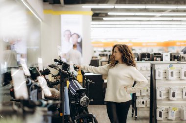 Modern bir elektronik mağazasında genç bir kadın odaklanma ve niyetle ürünlere göz atıyor. Görüntü teknolojiyi, alışverişi ve karar vermeyi ön plana çıkarıyor, davet edilen bir perakende satış ortamı sergiliyor.