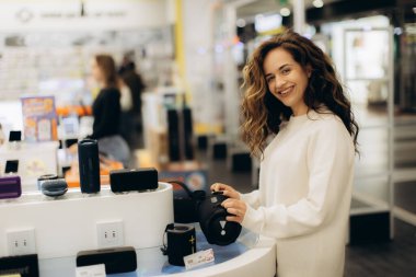 Neşeli genç bir kadın, bir teknoloji mağazasında hoparlörleri ve aletleri araştıran çeşitli elektronik cihazları araştırıyor. Yaşam tarzında modernlik ve tüketici elektroniği vurgulanıyor.