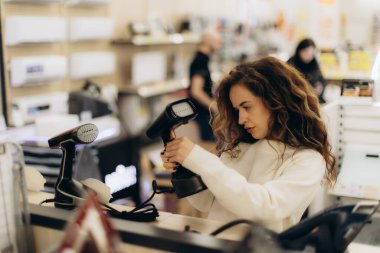 Bir mağazada modern elektronik aletlere göz atan, taşınabilir cihazlara odaklanan bir kadın. Özellikleri ve kalitesi hakkında detaylı bir değerlendirme yaparken aynı zamanda düşünceli bir alışveriş yapıyor..