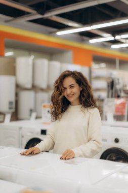 Neşeli bir kadın bir elektronik mağazasında duruyor, çeşitli modern aletlere bakıyor. Çevre, çağdaş yaşam tarzı ve tüketiciliği göstermektedir..