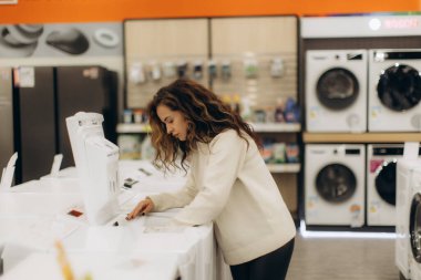 İyi aydınlatılmış bir elektronik mağazasında şık bir kadın satın almadan önce bir ev aletini dikkatle inceliyor. Ayar, modern ve pratik bir atmosfer yaratacak çeşitli cihazları içeriyor..