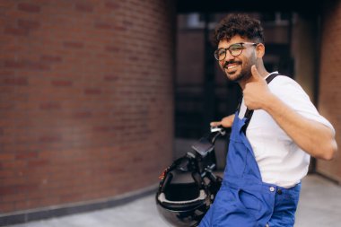 Mavi üniformalı bir kurye, motosikletini tutuyor ve şehir merkezinde bir duvarın yanında gülümserken başparmağını kaldırıyor.